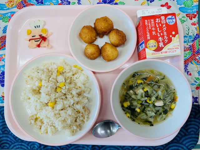 ピンクのトレーに牛乳、とうふドーナツ、コック帽をかぶった布製マスコット、ツナコーンピラフ、野菜スープと並べられている写真