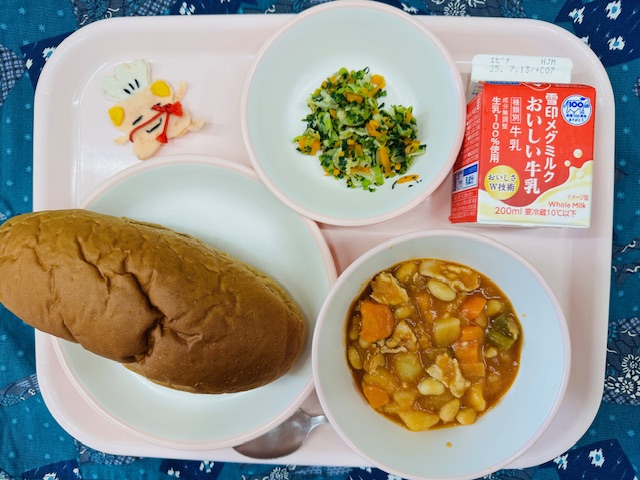 ピンクのトレーに牛乳、やさいソテー、コック帽をかぶった布製マスコット、くろパン、ポークビーンズと並べられている写真