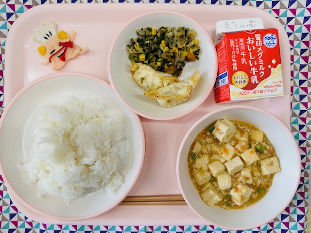 ピンクのトレーに牛乳、あげぎょうざとカラフルおひたし、コック帽をかぶった布製マスコット、ごはん、マーボーとうふと並べられている写真