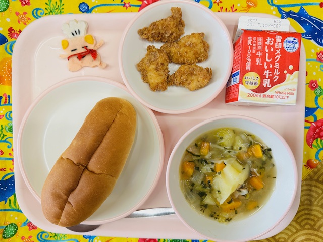 ピンクのトレーに牛乳、クリスピーチキン、コック帽をかぶった布製マスコット、ソフトフランスパン、やさいのスープ煮と並べられている写真