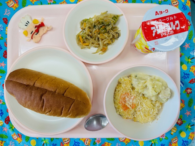 ピンクのトレーに発酵乳、しらたきサラダ、コック帽をかぶった布製マスコット、あじつきパン、ポテトグラタンと並べられている写真