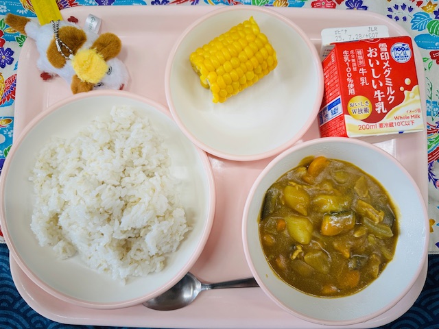 ピンクのトレーに牛乳、とうもろこし、犬のような布製マスコット、ごはん、なつやさいのカレーと並べられている写真