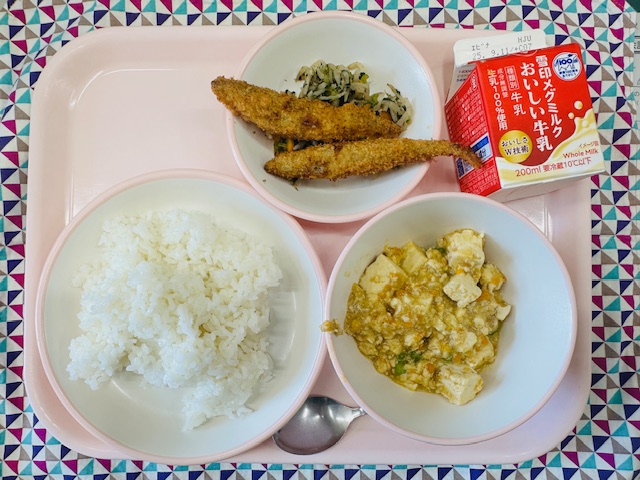 ピンクのトレーに牛乳、ししゃもフライともやしのおかかあえ、ごはん、マーボーどうふと並べられている写真