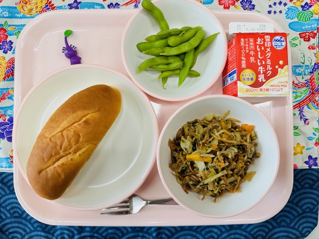 ピンクのトレーに牛乳、えだまめ、紫色の布製マスコット、ミルクパン、ソースやきそばと並べられている写真