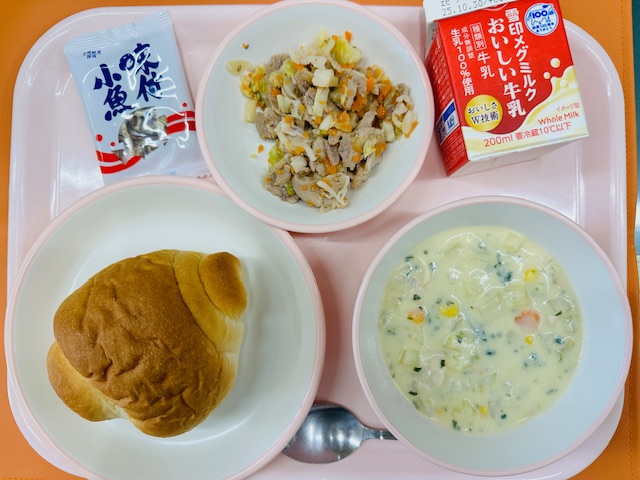 ピンクのトレーに牛乳、コーンのミルクスープ、ロールパン、味付き小魚、ゆでぶたサラダと並べられている写真