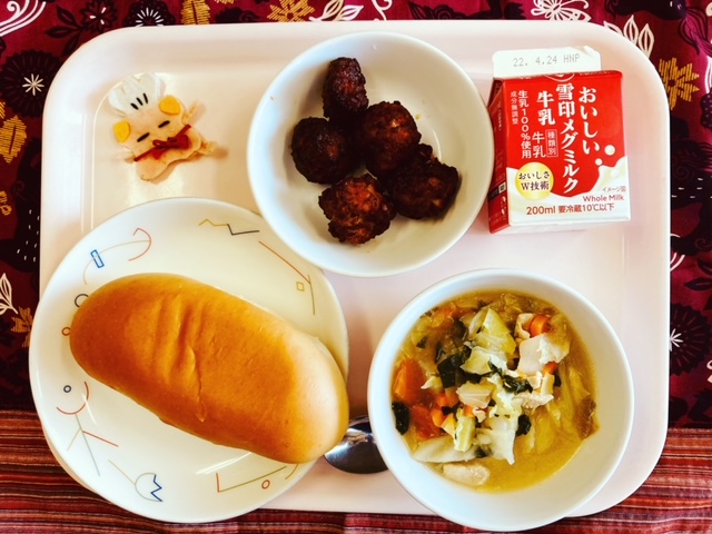 ピンクのトレーに給食が右上から反時計回りで牛乳、ビーンズミートボール、コック帽をかぶった布製マスコット、あじつけパン、野菜のスープ煮と並べられている写真