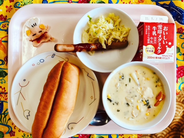 ピンクのトレーに給食が右上から反時計回りで牛乳、ウィンナー、キャベツ、コック帽をかぶった布製マスコット、パン、コーンシチューと並べられている写真