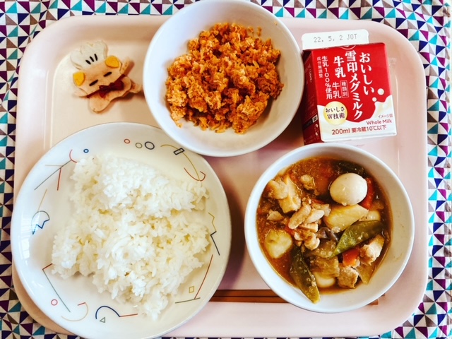 ピンクのトレーに給食が右上から反時計回りで牛乳、さけそぼろ、コック帽をかぶった布製マスコット、ごはん、春野菜のうま煮と並べられている写真