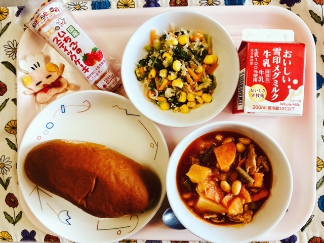 ピンクのトレーに給食が右上から反時計回りで牛乳、しらたきソテー、いちごのスティックケーキ（米粉）、コック帽をかぶった布製マスコット、くろパン、ポークビーンズと並べられている写真