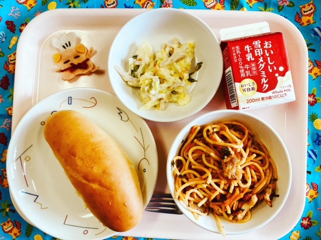 ピンクのトレーに給食が右上から反時計回りで牛乳、ポテトサラダ、コック帽をかぶった布製マスコット、ミルクパン、スパゲティペスカトーレと並べられている写真