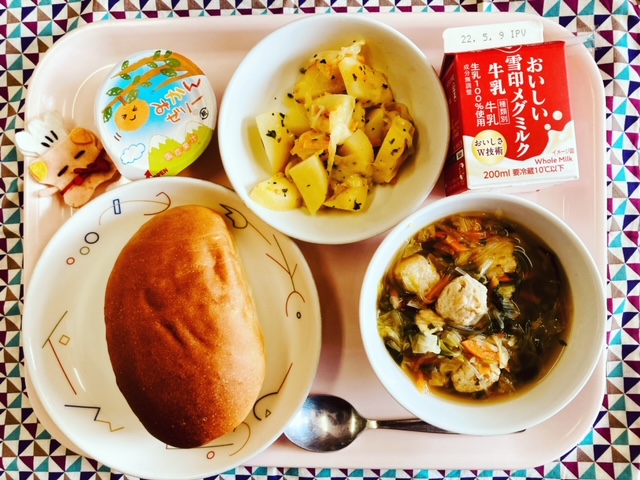 ピンクのトレーに給食が右上から反時計回りで牛乳、じゃがいものチーズ焼き、みかんゼリー、コック帽をかぶった布製マスコット、ソフトフランスパン、とり団子とキャベツのスープと並べられている写真