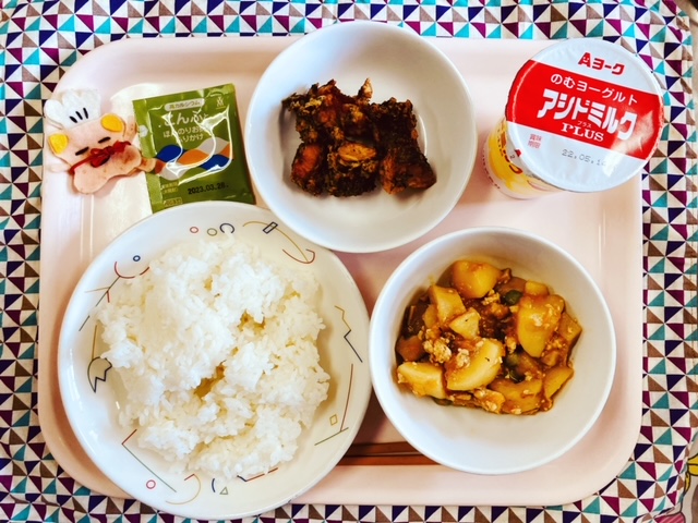 ピンクのトレーに給食が右上から反時計回りで発酵乳（いちごあじ）、魚のみなと揚げ、ふりかけ、コック帽をかぶった布製マスコット、ごはん、じゃがいものそぼろ煮と並べられている写真