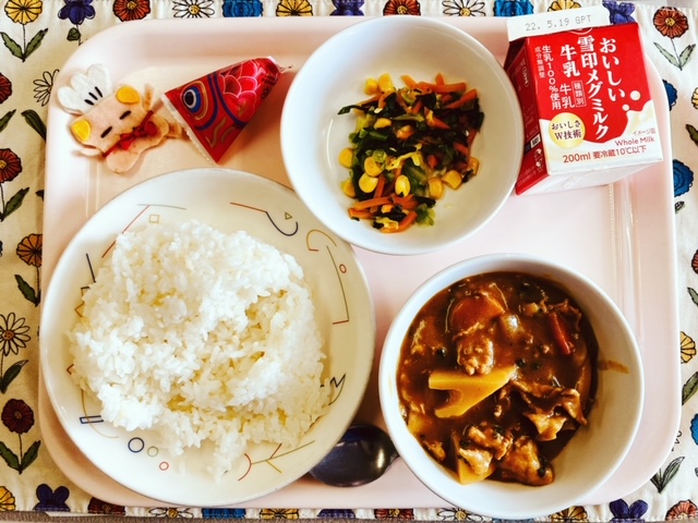 ピンクのトレーに給食が右上から反時計回りで牛乳、野菜ソテー、卵ぼーろ、コック帽をかぶった布製マスコット、ごはん、カレーライスのルーと並べられている写真