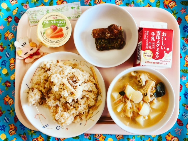 ピンクのトレーに給食が右上から反時計回りで牛乳、ちくわの磯部揚げ、フルーツキャロットゼリー、コック帽をかぶった布製マスコット、豚肉とタケノコの炊き込みご飯、みそ汁と並べられている写真