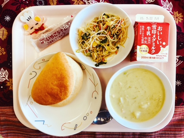 ピンクのトレーに給食が右上から反時計回りで牛乳、ビーフンソテー、いちごジャム、コック帽をかぶった布製マスコット、パン、グリーンシチューと並べられている写真