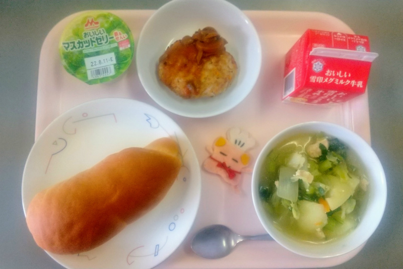 ピンクのトレーに給食が右上から反時計回りで牛乳、ハンバーグのデミグラスソースかけ、マスカットゼリー、ソフトフランスパン、コック帽をかぶった布製マスコット、やさいのスープ煮と並べられている写真