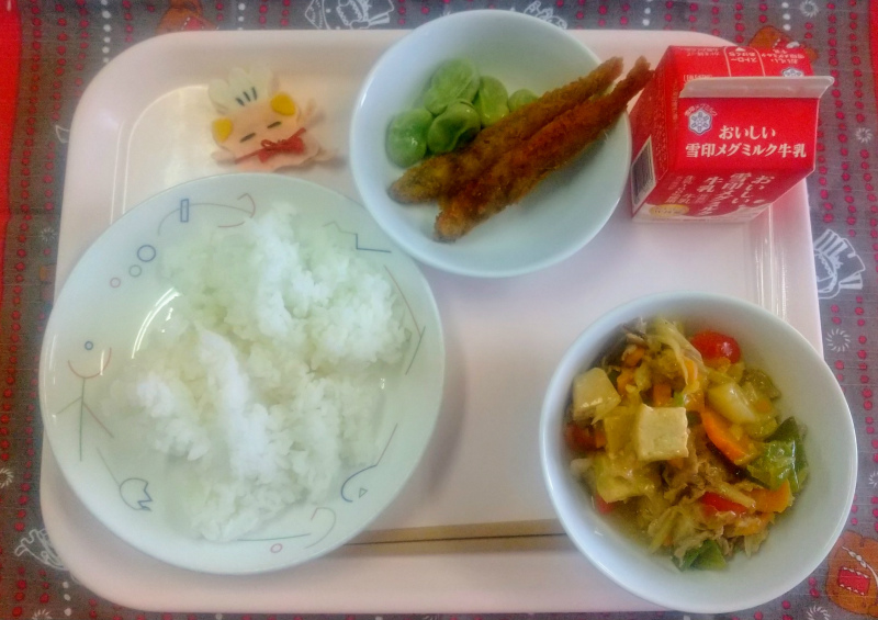 ピンクのトレーに給食が右上から反時計回りで牛乳、ししゃもフライとそらまめ、コック帽をかぶった布製マスコット、ごはん、ジャージャンとうふと並べられている写真