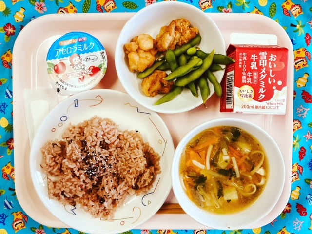 ピンクのトレーに給食が右上から反時計回りで牛乳、えだまめととりのから揚げ、アセロラゼリー、ごま塩がかかった赤飯、あんかけ汁と並べられている写真