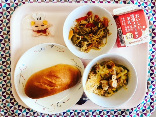 ピンクのトレーに給食が右上から反時計回りで牛乳、切り干し大根のサラダ、コック帽をかぶった布製マスコット、ミルクパン、ウィンナービーンズグラタンと並べられている写真