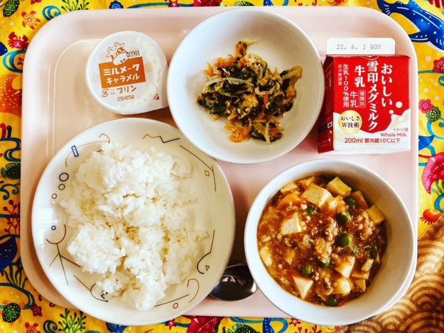 ピンクのトレーに給食が右上から反時計回りで牛乳、鶏肉と野菜のナムル、ミルメークキャラメルプリン、ごはん、マーボーどうふと並べられている写真