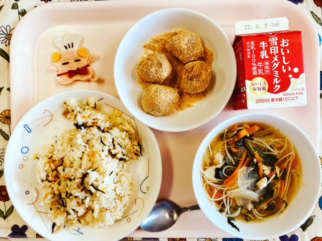 ピンクのトレーに給食が右上から反時計回りで牛乳、きなこドーナツ、コック帽をかぶった布製マスコット、ひじきピラフ、ビーフンスープと並べられている写真