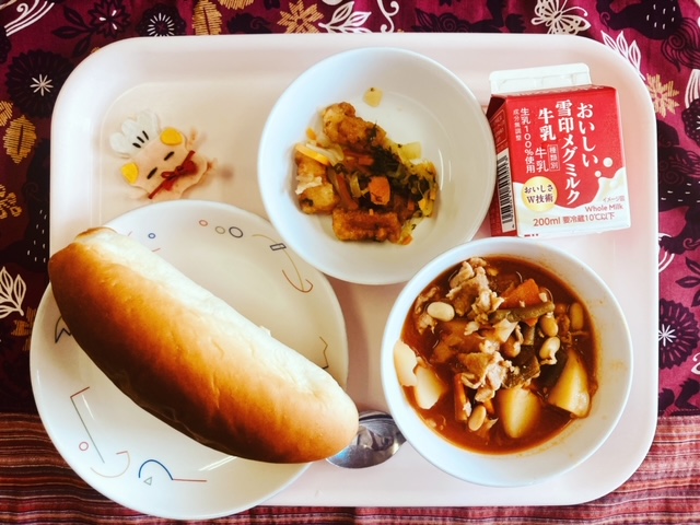 ピンクのトレーに給食が右上から反時計回りで牛乳、いかのマリネ、コック帽をかぶった布製マスコット、あじつけパン、ポークビーンズと並べられている写真