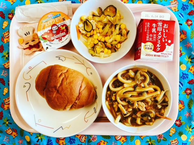 ピンクのトレーに給食が右上から反時計回りで牛乳、コーンサラダ、アセロラゼリー、コック帽をかぶった布製マスコット、ロールパン、カレーうどんと並べられている写真