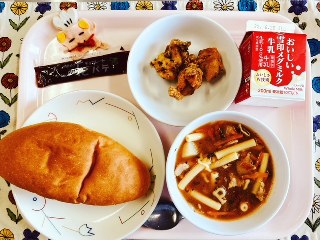 ピンクのトレーに給食が右上から反時計回りで牛乳、とりにくのハーブ揚げ、コック帽をかぶった布製マスコット、チョコパテ、コッペパン、ミネストローネと並べられている写真