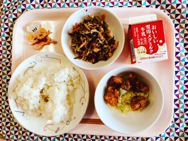 ピンクのトレーに給食が右上から反時計回りで牛乳、切り干し大根のミルク煮、コック帽をかぶった布製マスコット、グリーンピースごはん、とんかつ、キャベツソテーと並べられている写真