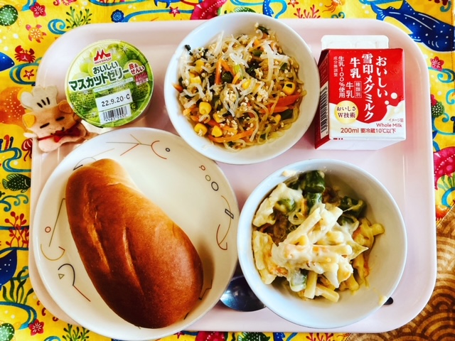 ピンクのトレーに給食が右上から反時計回りで牛乳、しらたきソテー、マスカットゼリー、コック帽をかぶった布製マスコット、ミルクパン、アスパラとうずらたまごのグラタンと並べられている写真