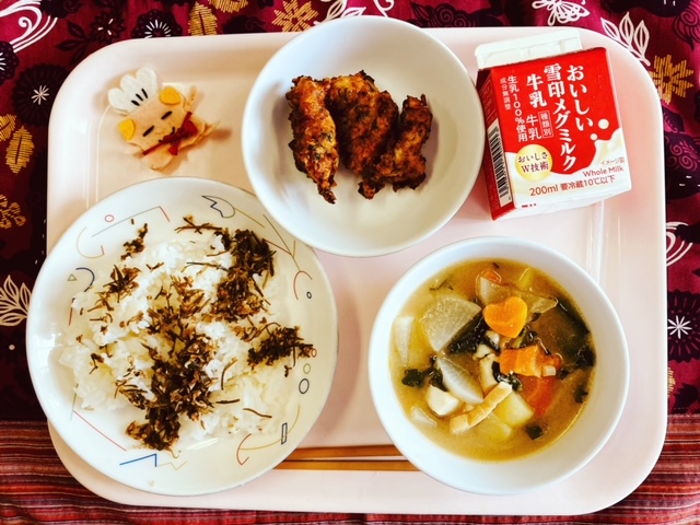 ピンクのトレーに給食が右上から反時計回りで牛乳、豚肉の衣揚げ、コック帽をかぶった布製マスコット、うめこんぶが載せられたごはん、みそけんちん汁と並べられている写真