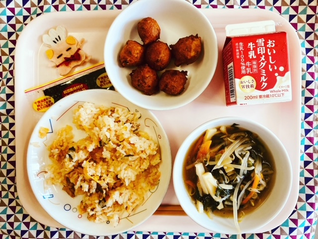 ピンクのトレーに給食が右上から反時計回りで牛乳、パンプキンボール、コック帽をかぶった布製マスコット、サラダのり、キムチごはん、ごもくスープと並べられている写真