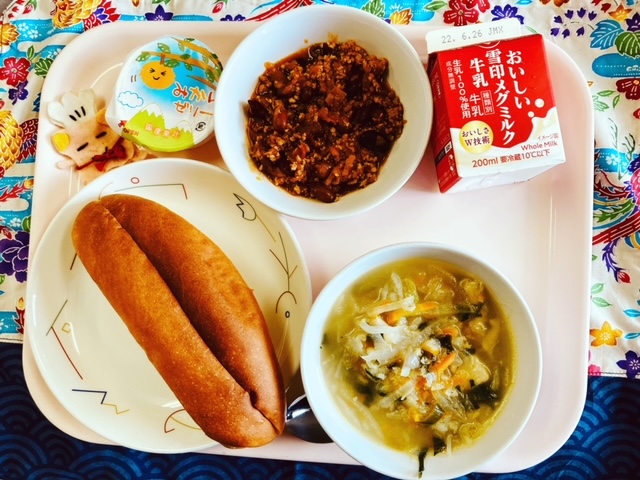 ピンクのトレーに給食が右上から反時計回りで牛乳、チリコンカンサンドの具、みかんゼリー、コック帽をかぶった布製マスコット、パン、スコッチブロスと並べられている写真