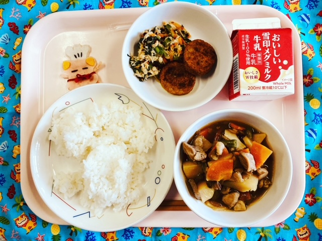 ピンクのトレーに給食が右上から反時計回りで牛乳、ごまあえ、しろごまつくね、コック帽をかぶった布製マスコット、ごはん、筑前煮と並べられている写真