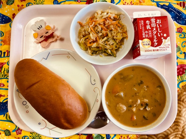 ピンクのトレーに給食が右上から反時計回りで牛乳、ハリハリサラダ、コック帽をかぶった布製マスコット、あじつけパン、カレーシチューと並べられている写真