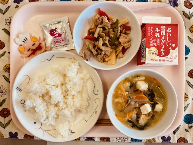 ピンクのトレーに給食が右上から反時計回りで牛乳、豚肉とピーマンの炒め物、うめじそふりかけ、コック帽をかぶった布製マスコット、ごはん、はるさめスープと並べられている写真