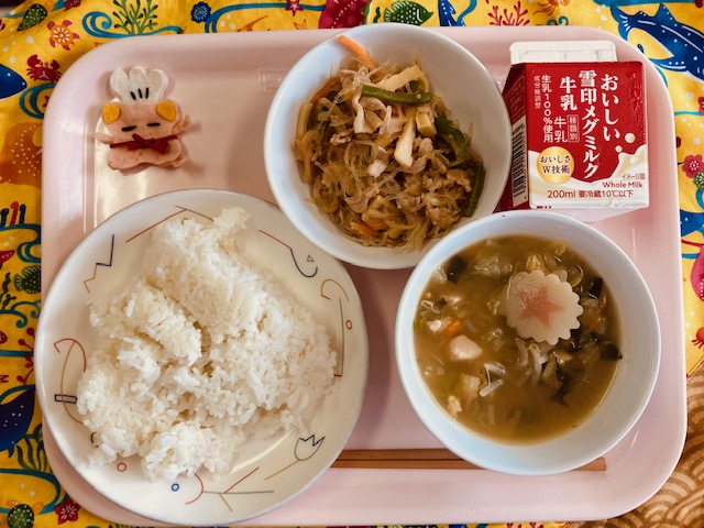 ピンクのトレーに給食が右上から反時計回りで牛乳、チャプチェ、コック帽をかぶった布製マスコット、 ごはん、トックスープと並べられている写真