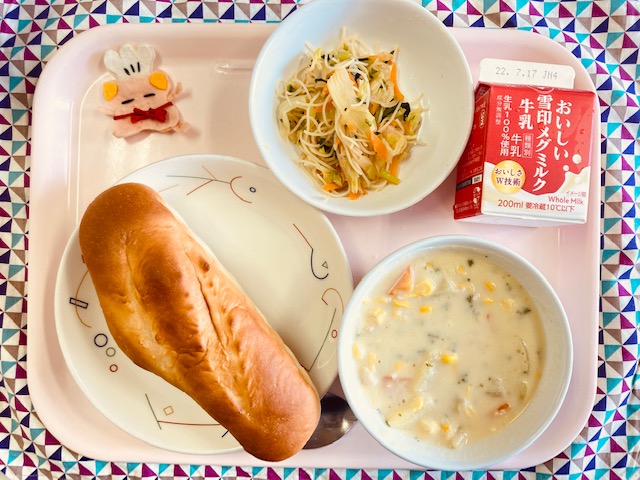 ピンクのトレーに給食が右上から反時計回りで牛乳、ビーフンソテー、コック帽をかぶった布製マスコット、ミルクパン、コーンシチューと並べられている写真