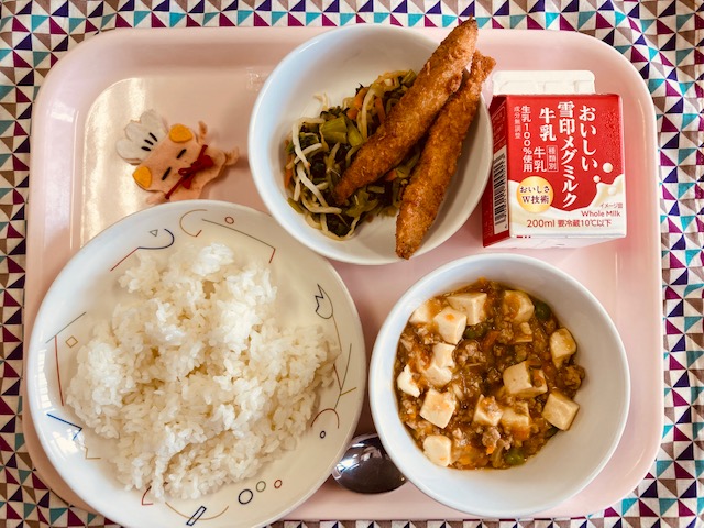 ピンクのトレーに給食が右上から反時計回りで牛乳、ナムル、ししゃもフライ、コック帽をかぶった布製マスコット、ごはん、マーボーどうふと並べられている写真