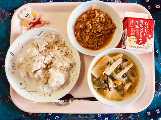 ピンクのトレーに給食が右上から反時計回りで牛乳、アジアンライスの具、コック帽をかぶった布製マスコット、アジアンライス、五目ワンタンスープと並べられている写真