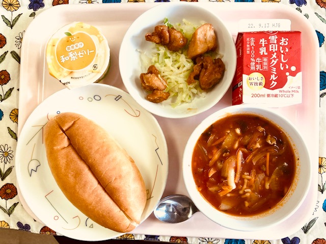 ピンクのトレーに給食が右上から反時計回りで牛乳、野菜、照り焼きチキン、和梨ゼリー、パン、ミネストローネと並べられている写真