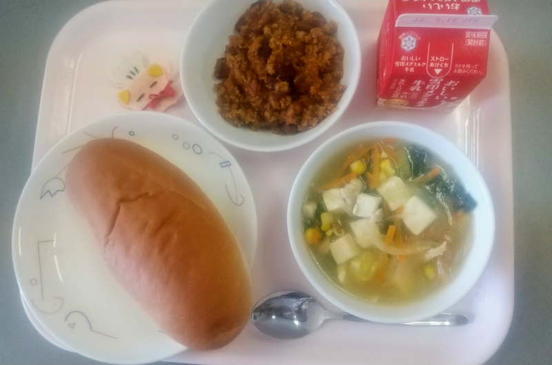 ピンクのトレーに給食が右上から反時計回りで牛乳、チリコンカン、コック帽をかぶった布製マスコット、あじつけパン、コーン中華スープと並べられている写真