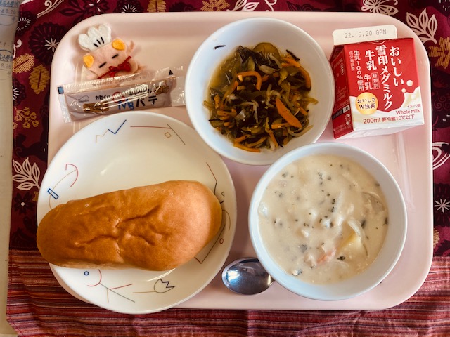 ピンクのトレーに給食が右上から反時計回りで牛乳、海藻サラダ、コック帽をかぶった布製マスコット、キャラメルパテ、ソフトフランスパン、まめシチューと並べられている写真