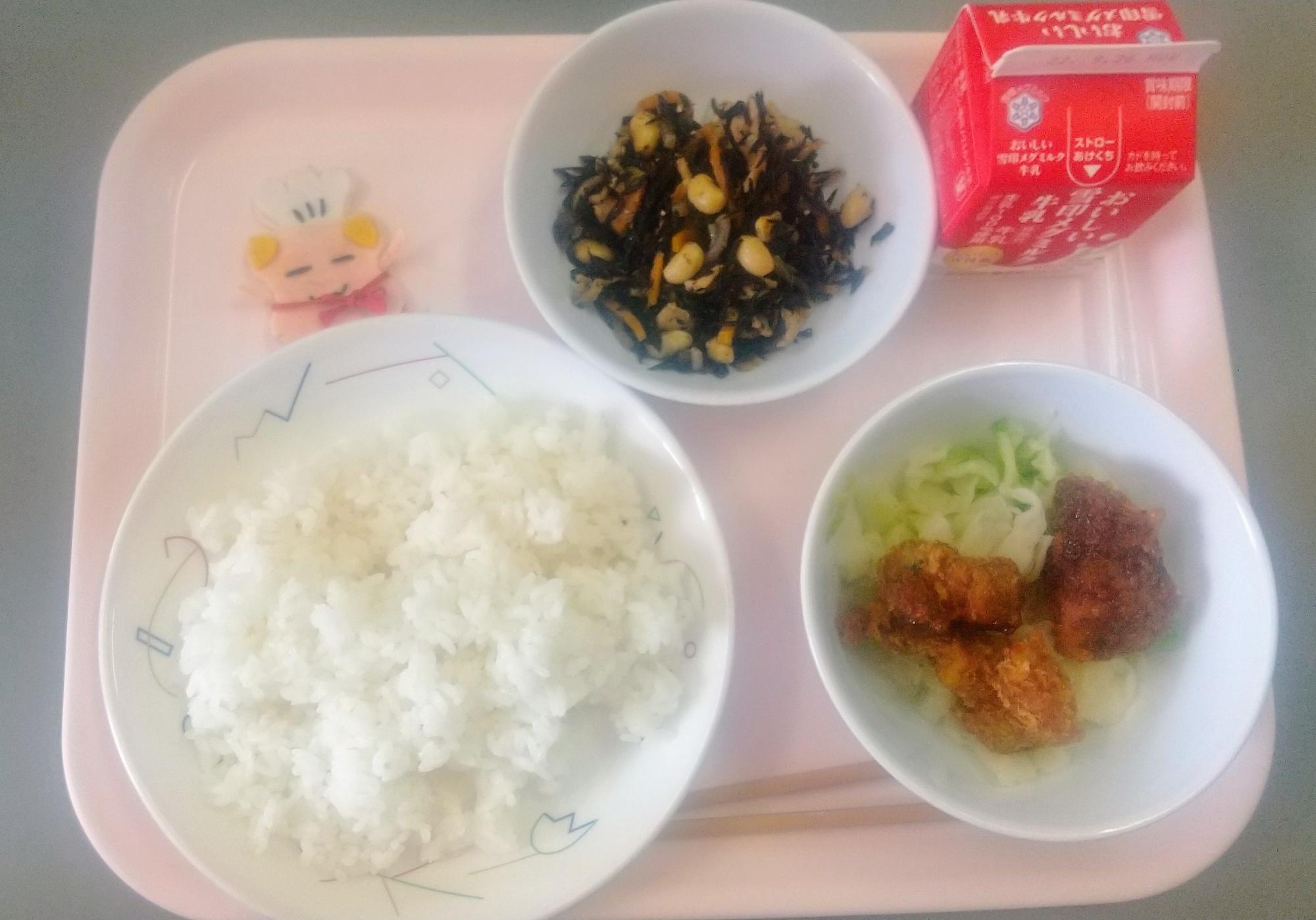 ピンクのトレーに給食が右上から反時計回りで牛乳、ひじきのいりに、コック帽をかぶった布製マスコット、ごはん、キャベツソテー、とんかつと並べられている写真