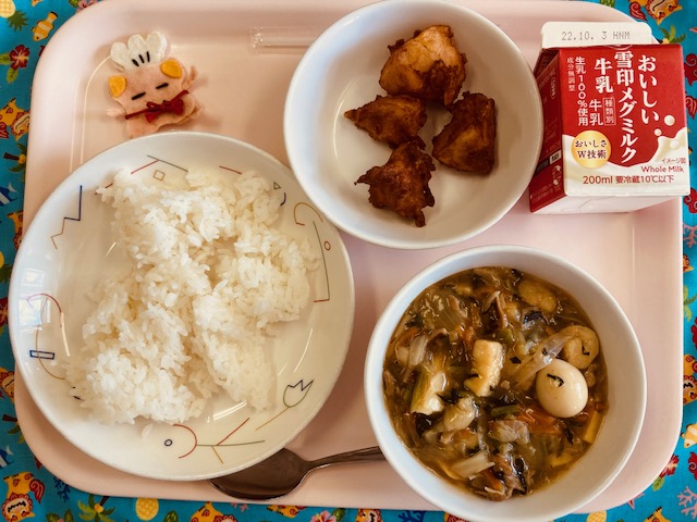 ピンクのトレーに給食が右上から反時計回りで牛乳、じゃがじゃが、コック帽をかぶった布製マスコット、ごはん、中華丼のあんと並べられている写真