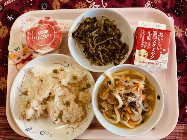 ピンクのトレーに給食が右上から反時計回りで牛乳、いそかあえ、りんごゼリー、コック帽をかぶった布製マスコット、かしわずし、にくうどんと並べられている写真