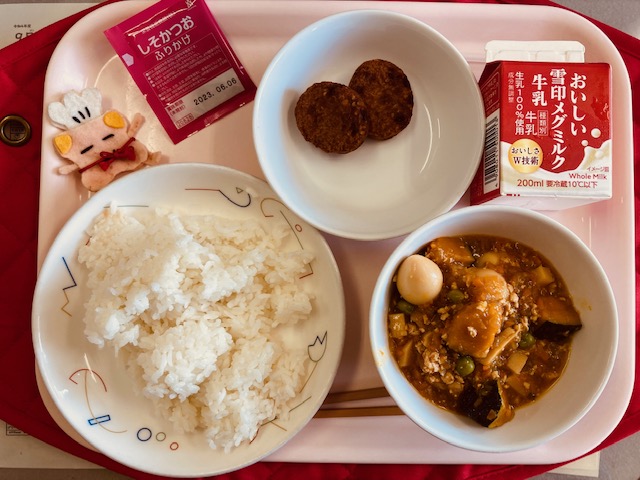 ピンクのトレーに給食が右上から反時計回りで牛乳、しろごまつくね、しそかつおふりかけ、コック帽をかぶった布製マスコット、ごはん、かぼちゃのそぼろ煮と並べられている写真