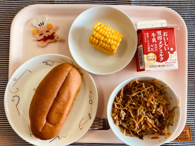 ピンクのトレーに給食が右上から反時計回りで牛乳、とうもろこし、コック帽をかぶった布製マスコット、パン、焼きそばと並べられている写真
