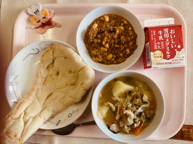 ピンクのトレーに給食が右上から反時計回りで牛乳、キーマカレー、コック帽をかぶった布製マスコット、ナン、野菜のスープ煮と並べられている写真