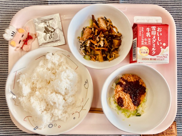 ピンクのトレーに給食が右上から反時計回りで牛乳、切り干し大根の炒め煮、ふりかけ、コック帽をかぶった布製マスコット、ごはん、キャベツソテー、メンチカツと並べられている写真